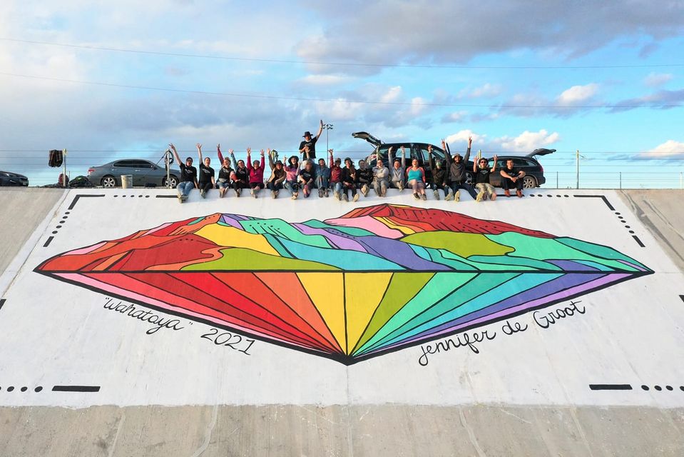A large group of people sitting in a line on white levee wall with their arms raised in celebration. In the foreground, a large colorful geometric mural is displayed on the wall, featuring vibrant rainbow-colored mountain landscapes and radiating lines in a fan or sunrise pattern. The sky above shows blue skies with white and gray clouds.